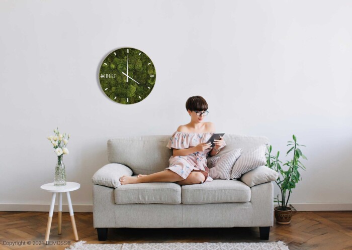 Eine Frau sitzt auf einer Couch mit einem Buch in der Hand und einer Uhr an der Wand hinter ihr, moosbild, mooswand, moos pflanzen, moos, moos deko, moos art